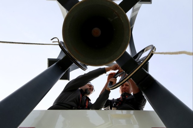 Chièvres Air Base Bell Tower: Two men stand in the bucket of a cherry picker lift while holding a wheel connected to a bell tower; photo taken from directly below. 