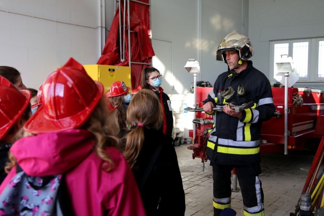 A firefighter stands indoors and talks to children wearing red fire fighter helmets about equipment used during a fire. 