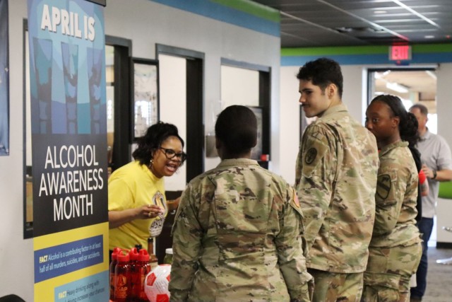 A woman in a yellow t-shirt speaks to three people wearing Army operational combat uniforms. 