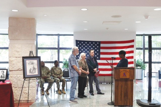 One person standing behind a wooden podium looks reads off a certificate while three people stand in a row with two people sitting behind them off slightly to the side. 