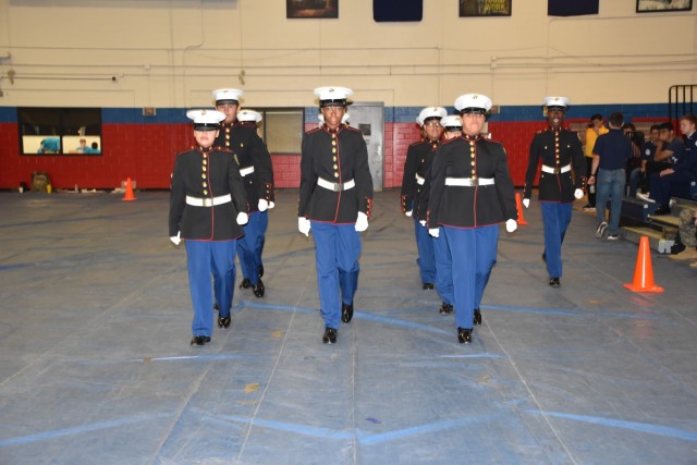 Ten people in U.S. Marine JROTC dress uniform march toward the camera on the floor of a gymnasium. 