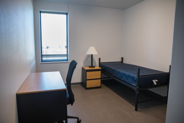 a room with a black twin bed frame, a dark blue mattress sitting on the bed fram, a wooden night stand, a lamp with a white shade, a wooden desk and an office chair. 