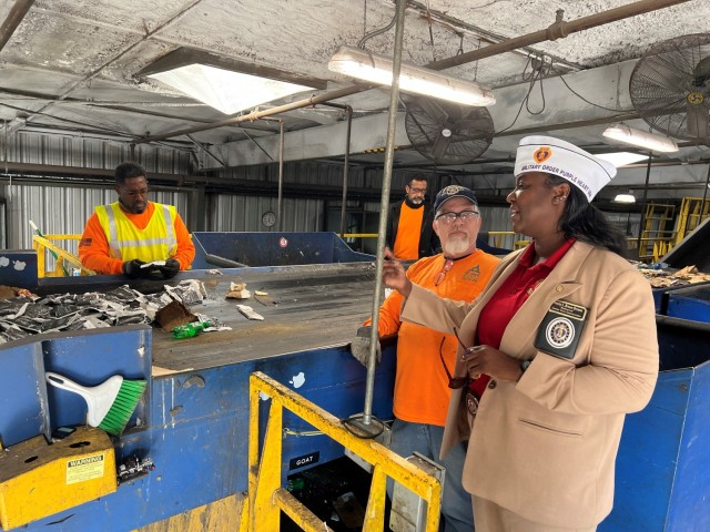 Two people sort through recycling on a conveyer belt while two other people converse on the other side of the belt. 