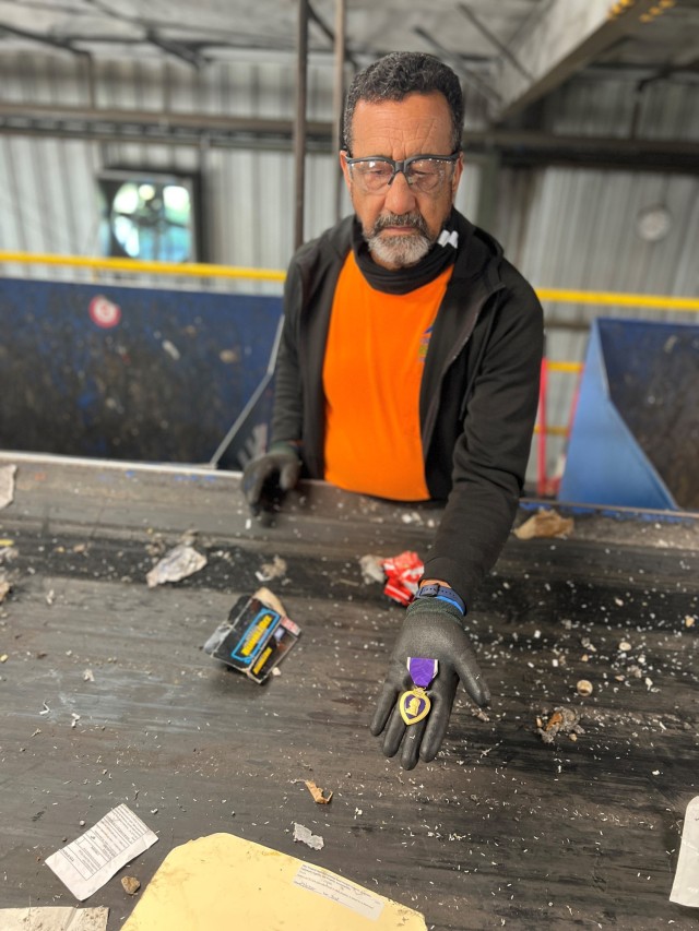A man standing holds one hand out with a gold medal in the shape of a heart and a purple ribbon.