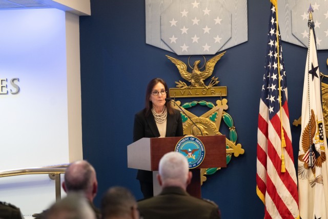 A woman standing behind a podium speaks to a crowd of people sitting. 
