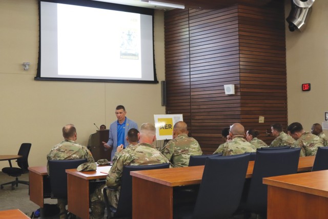 A man standing talk to people sitting in chairs and behind long tables. 