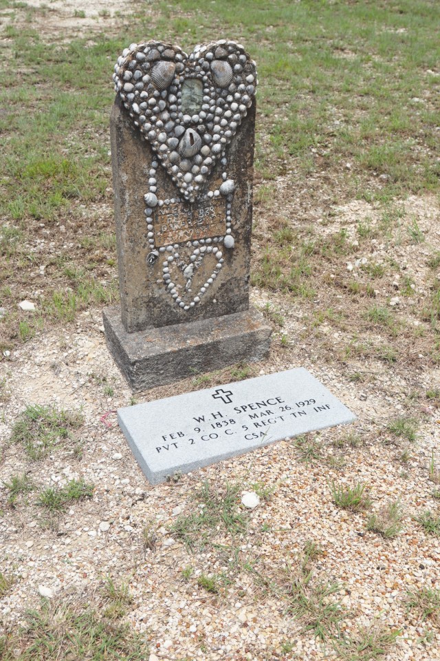 A headstone sporting a stone heart at the top encrusted with small stones and seashells sits in an open area of grass. 