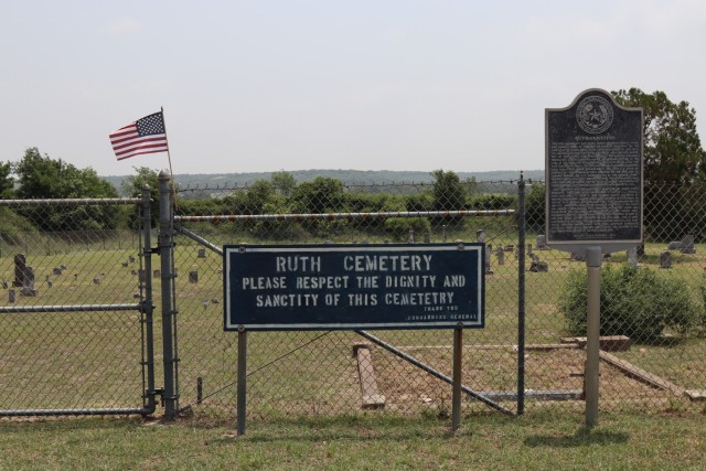 A metal gate that connects to a fence with barbed wire running on top sits behind a slightly shorter sign.