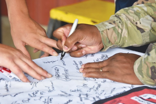A hand holding a permanent marker writes on a cloth banner while the other three hands hold the banner taut and flat. 