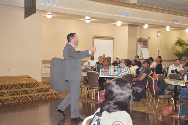 A man standing talks in front a crowd of people sitting at tables in a large room. 