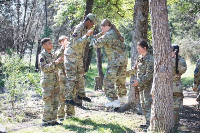 Two teenagers hold hands while steadying themselves on separate wires tied between two trees as other look on. 