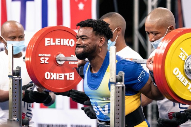 A man squats a metal bar resting on his shoulders with four weights on each side as three other men stand nearby.