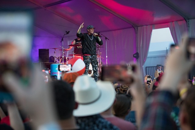 A man holds a microphone while on a stage as many people standing look on, some holding phones recording in the air.