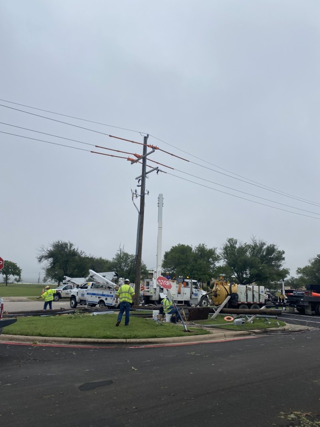 Three men in bright yellow shirts and construction hard hats look at a fallen, broken electrical pole in a patch of grass. 