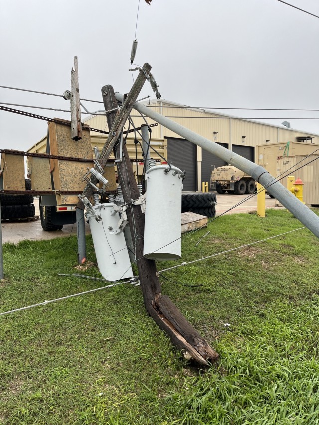 An electric pole lays on a patch of grass, it's wood snapped, broken and bent, the metal items attached to also in similar conditions.