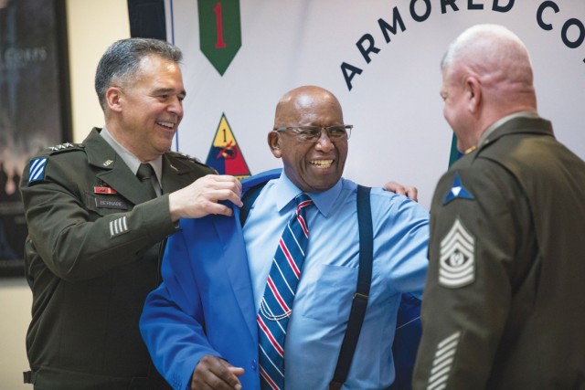 A man assists another man in putting on a blazer as they smile while a different man looks on. 