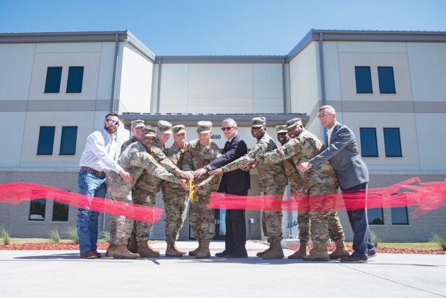Several people hold a giant pair of scissors and pose for a photo in front of a building as red ribbon falls to the ground.