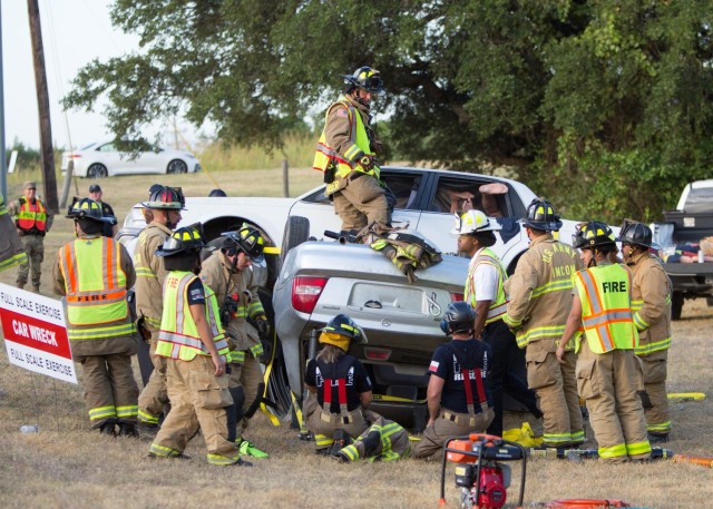 People in firemen uniform stand around an overturned car and another angled on its side.