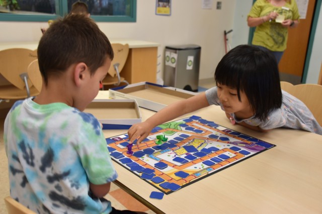 Two children look down at a board game on a table as one of the children moves a purple piece.