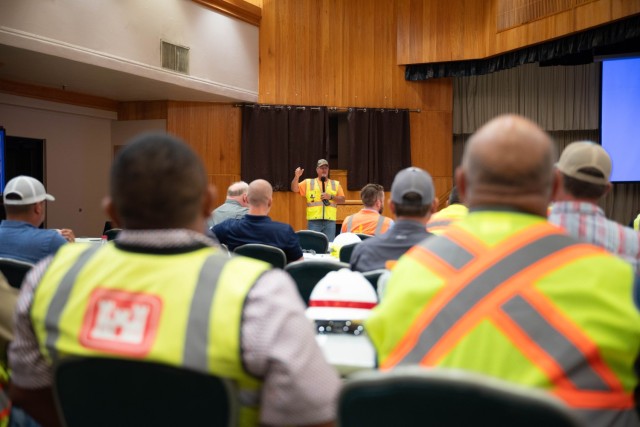 People sitting in chairs look on at a man standing and talking into a mic he is holding.