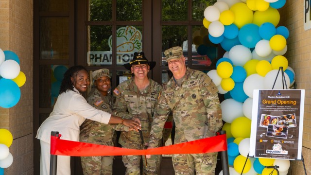 Four people pose for a picture behind two poles with red ribbon tied between them, the four people holding a giant pair of scissors to the ribbon.