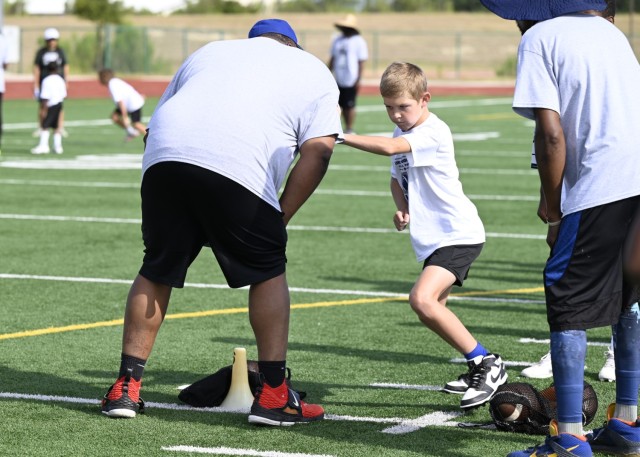 A young boy puts his left hand on a man's shoulder and prepares to run around the man, lifting his left foot, as another man looks on at them.