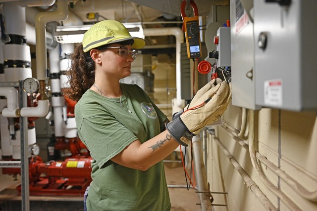 A women standing wears a hard hat and gloves while holding electric wires to an  electrical circuit.