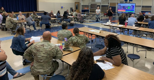 People sit at rows of table in large room. At the front of the room, two standing face the people sitting, one gesturing toward herself with her hands.