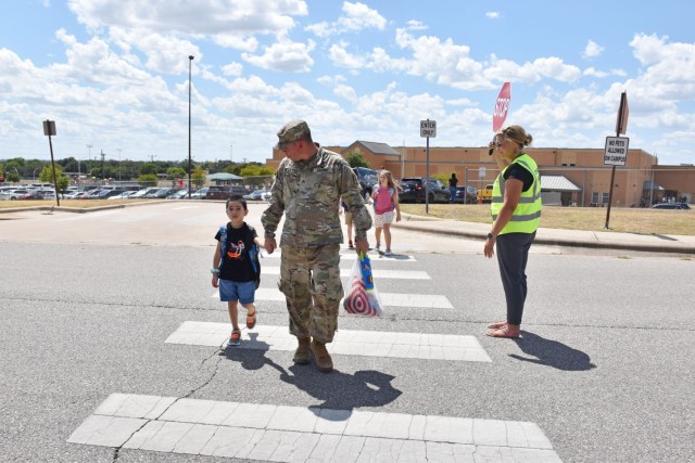 A man and a child hold hands as they cross a crosswalk while a woman in a bright, reflective yellow vest holds a red, octagonal sign that reads "STOP."