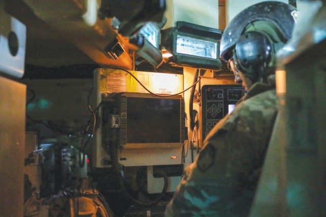 A man sitting and dressed in an operational combat pattern uniform, a black helmet and hearing protection, looks at an electronic screen in a small space.