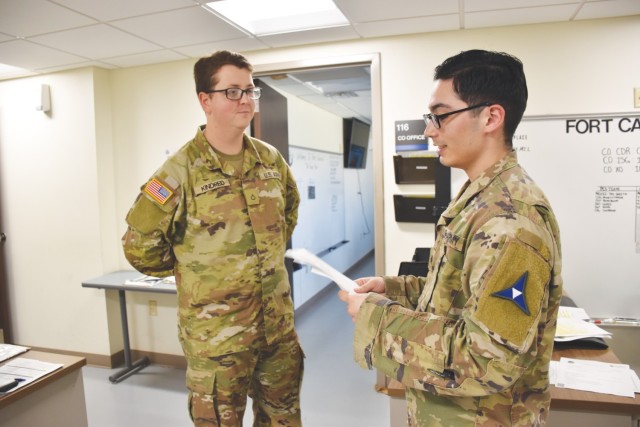 A man in glasses and dressed in and operational combat pattern uniform, or OCPs, speaks while looking at a paper held in his hands as another man dressed in OCPs and in glasses looks at him.