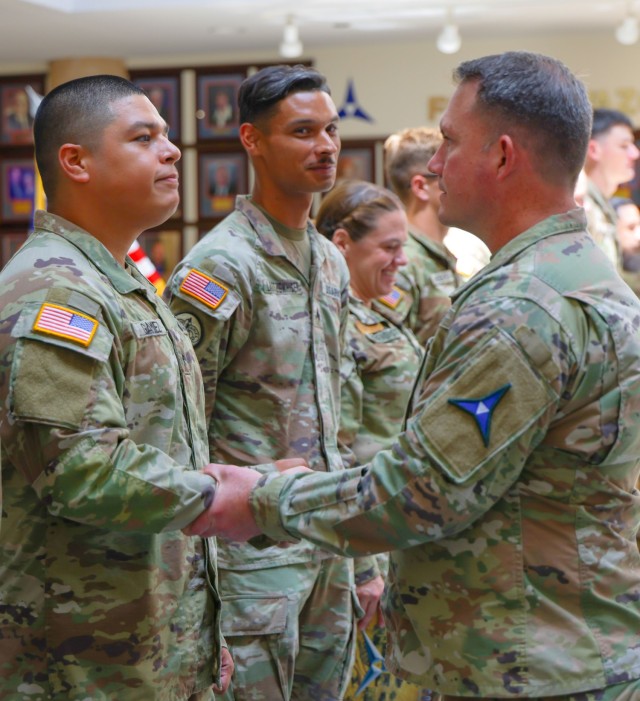 Two men dressed in operational combat pattern, or OCP, uniforms shake hands while standing as other people dressed in OCPs stand in a line side by side nearby.