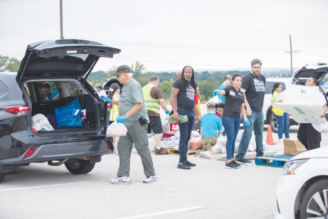 An older man moves toward an open car trunk holding a plastic bag, as other people look on.