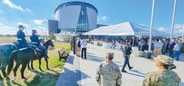 Tens of people sit and stand in front of a large, circular building, their bodies oriented toward the building.