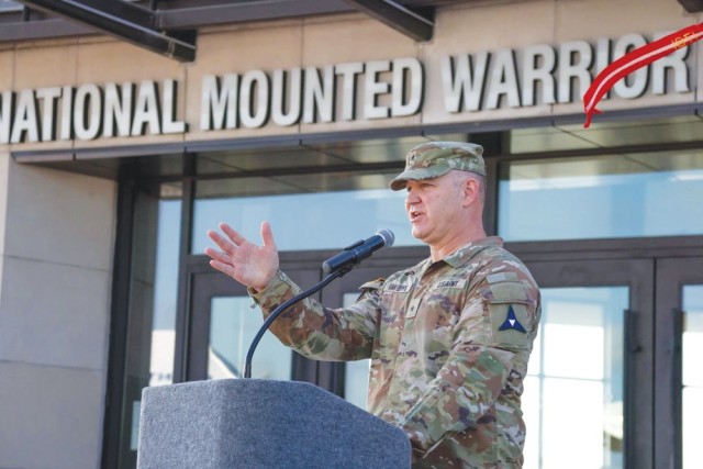 A man in an Army combat uniform standing behind a podium with a microphone attached to it speaks, gesturing with his right hand while doing so.