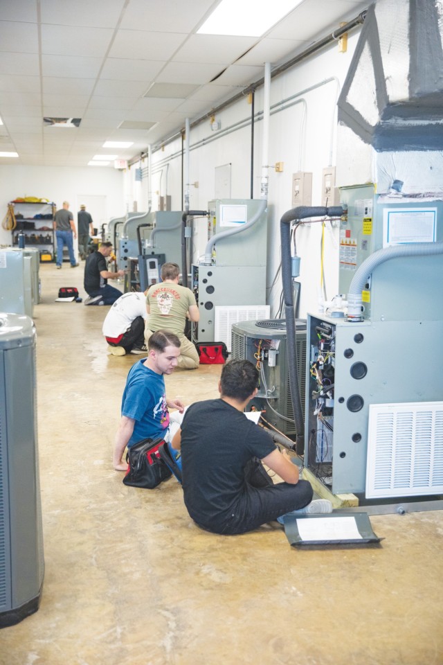 Several people sitting in pairs sit in front of air conditioning units with wires exposed.