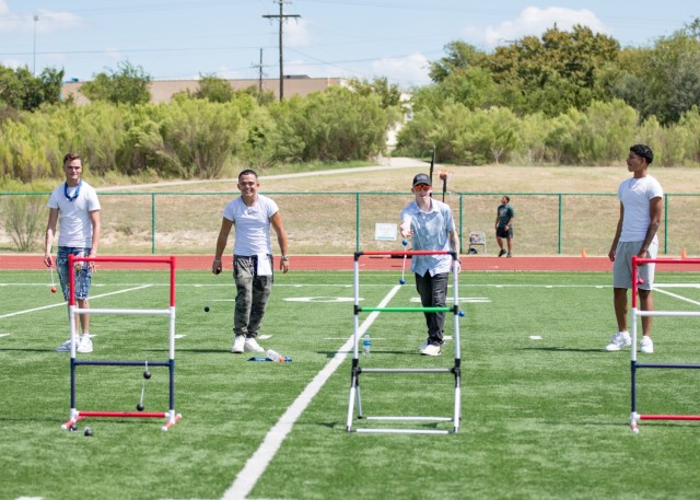 Four men stand on a turf field, flinging strings with two balls attached to the ends toward short, standing ladders. Far behind them, a man watches as a basketball goes into a basketball hoop.