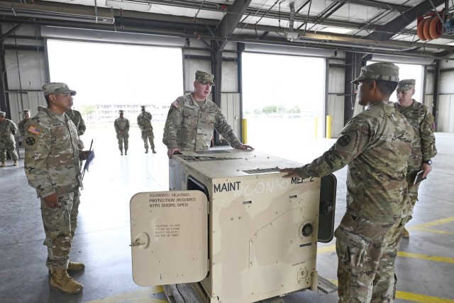 Four men dressed Army combat uniforms stand around a large, rectangular, metal piece of equipment sitting on a wooden pallet as others far behind stand and look at them.