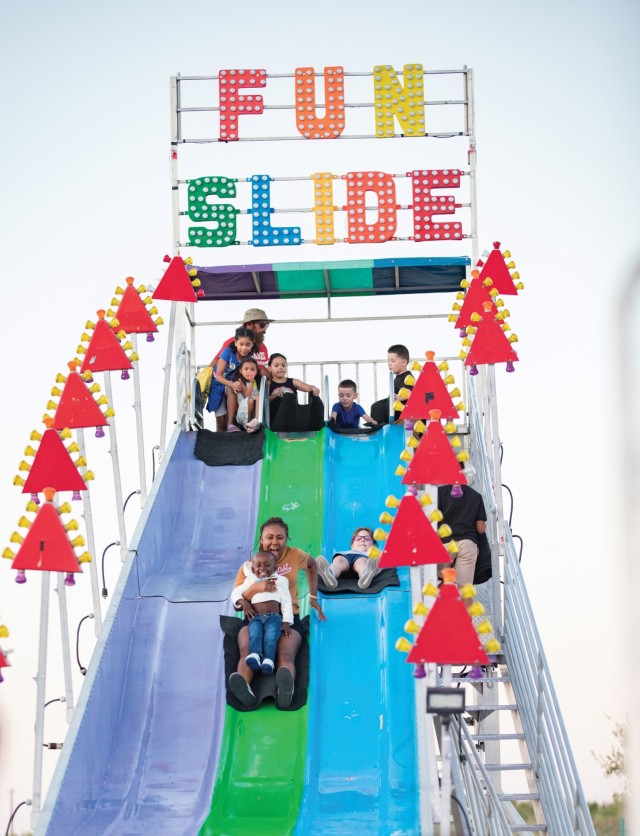 Several people atop long black mats slide down three slides connected together side by side.