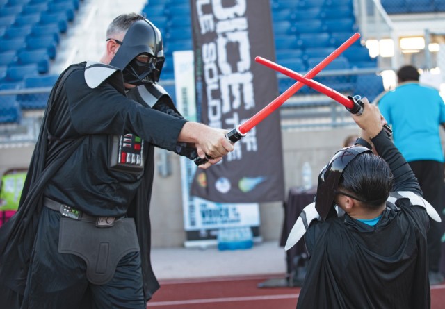 Two men fight with red replica lightsabers while wearing Darth Vader costumes.