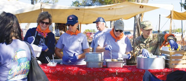 Four people stand side by side behind a red-clothed table with large, metal containers sitting on top.