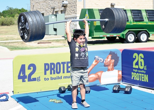 A young boy holds a fake barbell with weights over his head while standing tall.