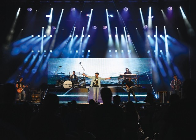 A man stands behind a mic stand with his arms spread wide on a stage, while other men around him play various instruments.