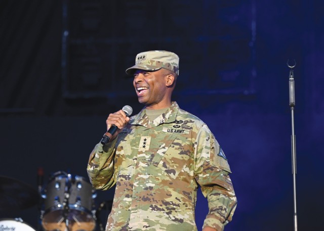 A man dressed in an Army combat uniform holds a microphone in his right hands while standing and smiling.