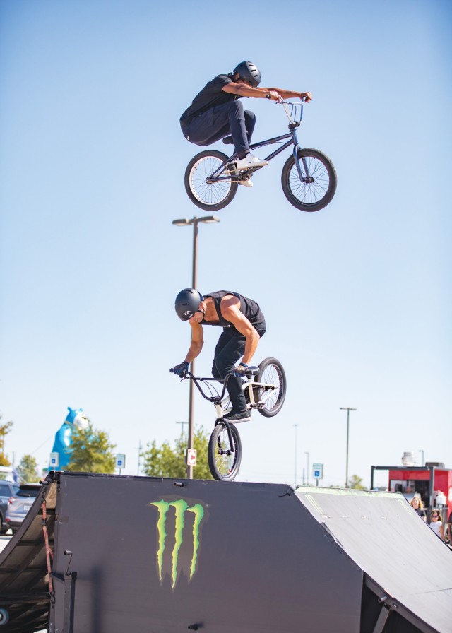 A man is in the air mounted on a bike. Straight below him, another man on a bike balances the bike on its front wheel.