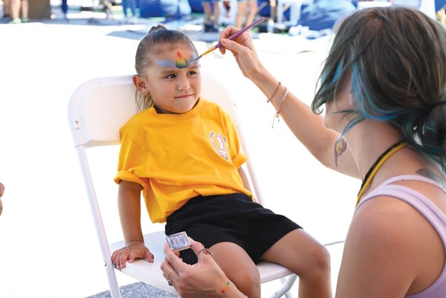 A young girl sits in a chair while a woman paints on the girl's forehead with a thin paint brush.