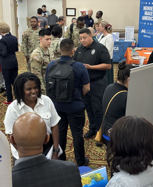 People stand looking at infographics and talking to people while sitting in front of a row of clothed-covered tables with freebies and informational papers sitting on top of them.