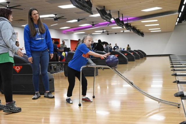 A young girl pushes a bowling ball off a bowling ramp into a lane as two adult women stand, looking on.