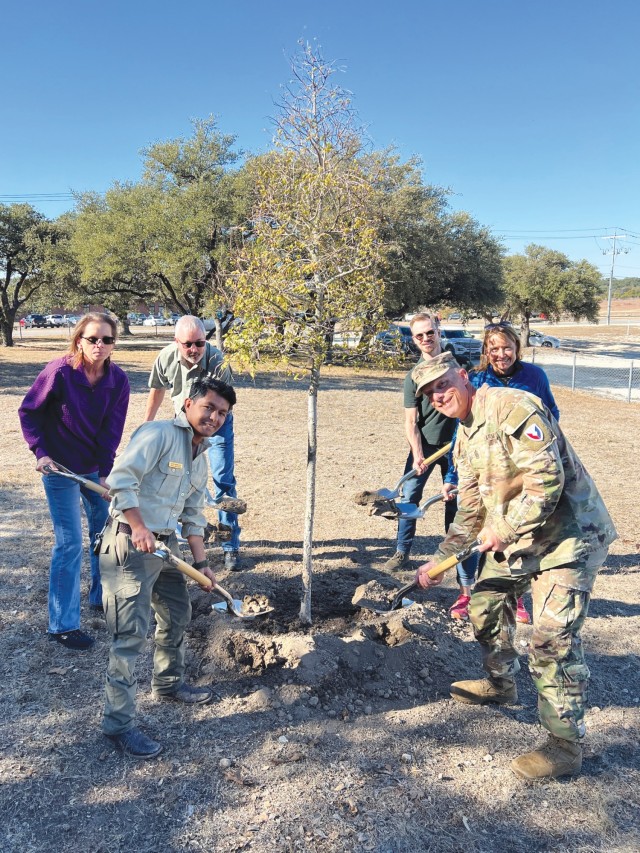 A group of people standing holding shovels with dirt in them pose for a photo around a tree.