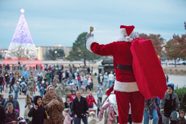 A man standing with his back to the camera with a beard white as snow dressed in a red suit with white accents and a christmas hat holds a bell in his left hand outstretched toward a crowd of people.
